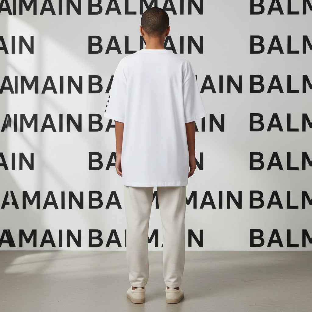 Noticed Atelier by Legzdiamond Balmain white oversized men's t-shirt viewed from the back against a branded backdrop.